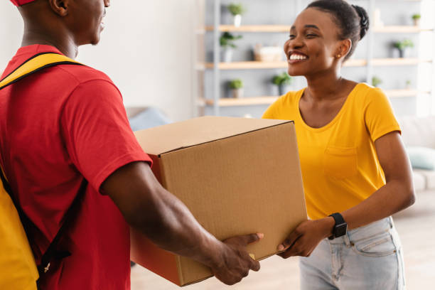 Delivery Concept. Happy African American Woman Receiving Big Box From Courier Man In Red Uniform Standing At Doors Of Her Home. Cropped Shot, Selective Focus On Paperboard Parcel.